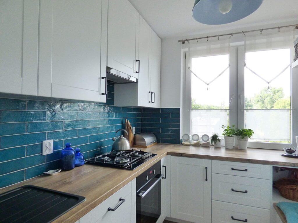 Na zdjęciu widzimy urokliwą kuchnię na wymiar, której serce stanowi harmonijna mieszanka bieli i intensywnego odcienia turkusowych kafelków. Meble kuchenne są utrzymane w jasnej kolorystyce, co optycznie powiększa przestrzeń i dodaje jej lekkości. Drewniane blaty doskonale kontrastują z nowoczesnymi, czarnymi uchwytami, tworząc wyważoną całość. Wnętrze jest świetnie doświetlone zarówno dzięki naturalnemu światłu płynącemu przez okno, jak i delikatnemu oświetleniu umieszczonemu pod szafkami. Dodatki takie jak drewniany młynek do pieprzu, elegancka czajnik na gaz oraz rośliny doniczkowe nadają kuchni domowy i ciepły klimat.Firma Aksent jako producent kuchni na wymiar specjalizuje się w tworzeniu przestrzeni dostosowanych do indywidualnych potrzeb każdego klienta. Ta kuchnia może stanowić przykład ich rzemiosła, gdzie dbałość o detale, jakość wykonania i funkcjonalność są na pierwszym planie. Tworząc spersonalizowane kuchnie, Aksent bierze pod uwagę nie tylko estetyczne aspekty, ale i codzienną wygodę użytkowania, łącząc je w unikatowe projekty, które idealnie wpasowują się w życie użytkowników. Kafelki dodają kuchni charakteru, a całość designu odzwierciedla współczesne trend w aranżacji wnętrz, pozostając przy tym czasowo i funkcjonalnie uniwersalna.